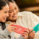 Two women hold the Switch Lite, one pink and the other green.
